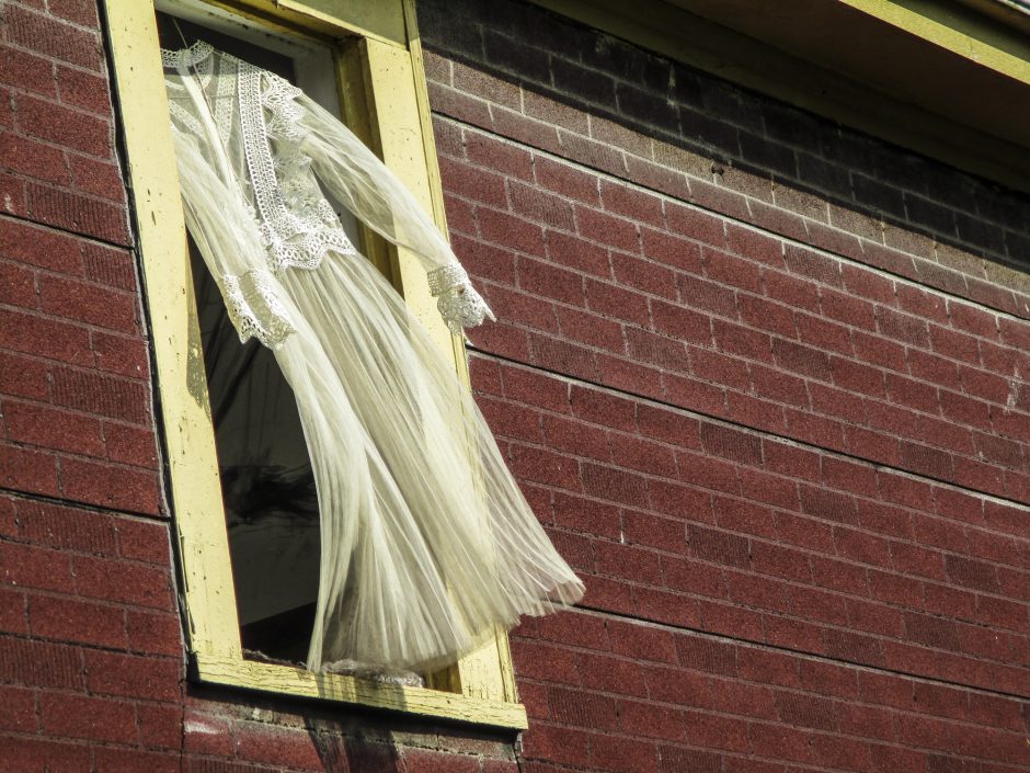 Image shows exterior of a building with red faux-brick siding. A window to the left of the image has a yellow frame and a yellow-white dress on a clothes hanger is billowing in the frame like a curtain.