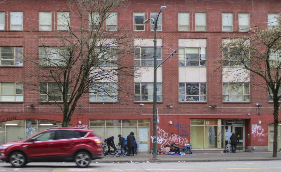 Exterior of a brick building in Vancouver's Downtown Eastside. Bricks have graffiti. People with carts and walkers stand, walk and sit in front of the building. A car is moving to the left, about to leave the frame, car wheels are blurry indicating movement.