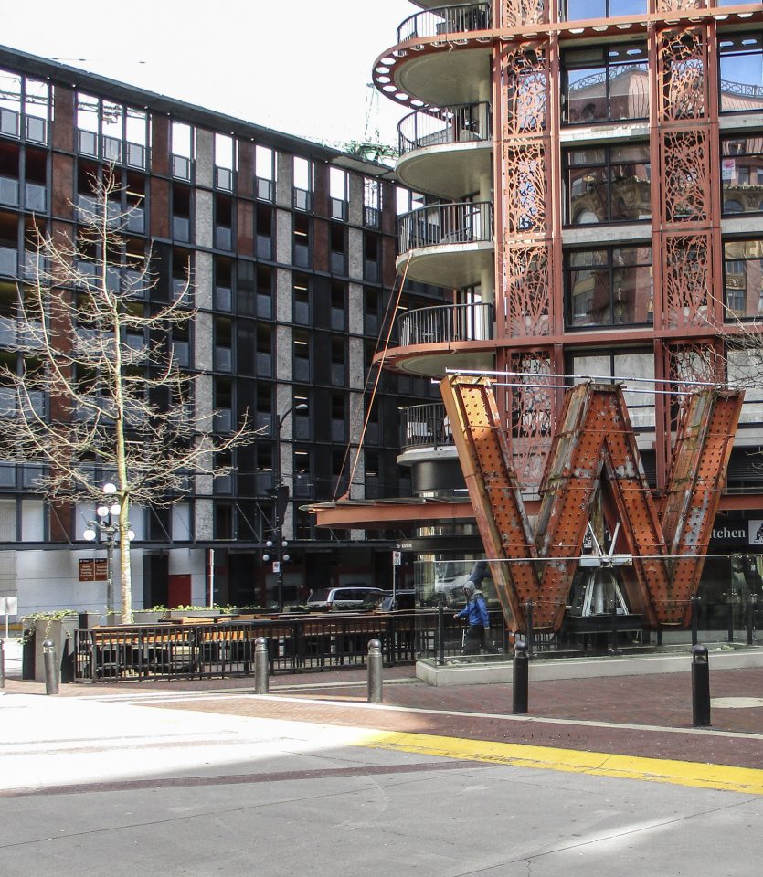 Image shows large "W" sign from the old Woodwards building. The sign is installed in front of the SFUWoodwards building that is now on the site. A man in a blue coat walks out from behind the W.