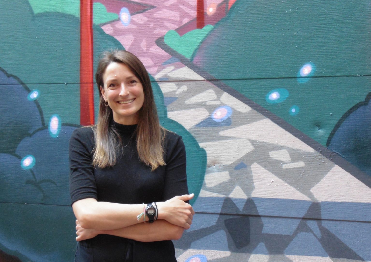Alina McKay, former graduate research assistant at the UBC Learning Exchange, stands and smiles with arms folded in front of the mural in the alley behind the Learning Exchange.