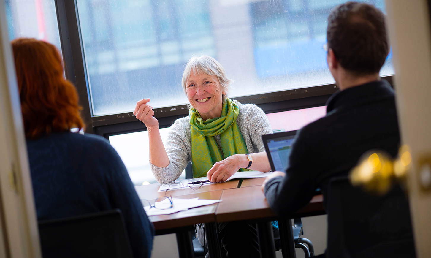 Angela Towle takes part in a meeting at the UBC Learning Exchange. She's smiling and wearing a lime green scarf over an off-white sweater and is gesturing to make a point as she speaks.